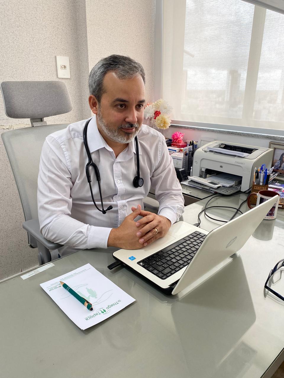 Dr. Thiago Tapioca, médico clínico em Feira de Santana, em seu consultório, sentado à mesa com estetoscópio